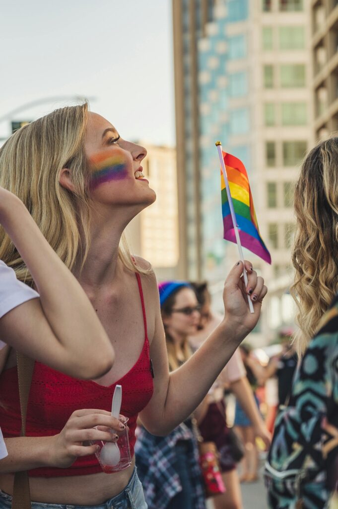 Smiling woman with rainbow face paint waving a flag at a lively Pride parade.
